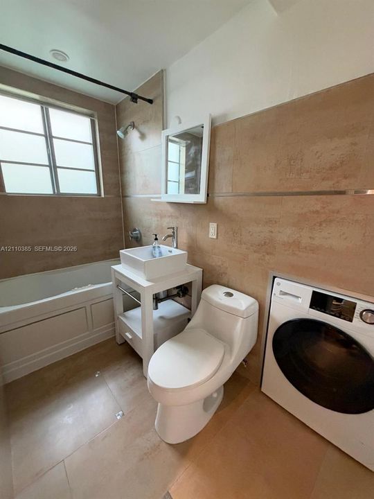 Bathroom featuring large-format wall and floor tile, a vessel sink on a white open-frame vanity, a built-in soaking tub, and an integrated laundry appliance