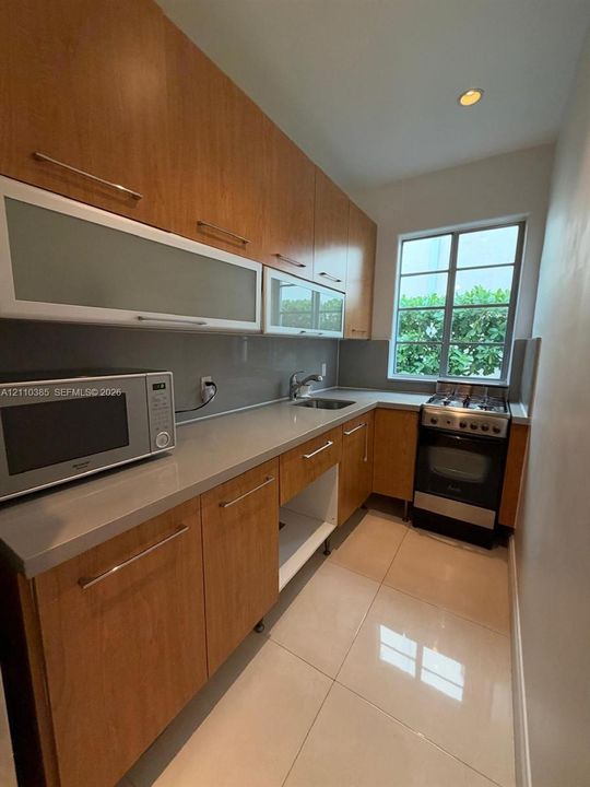 Kitchen featuring wood-finish cabinetry, neutral solid-surface countertops, large-format tile flooring, a stainless steel gas range, and upper cabinets with frosted glass panels