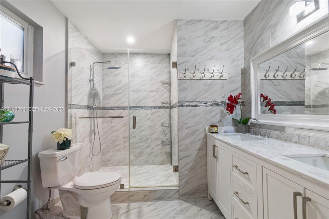 Bathroom featuring stone-finish tiling, walk-in glass-enclosed shower with rainfall showerhead, dual-sink vanity with light-tone countertop, and wall-mounted mirror