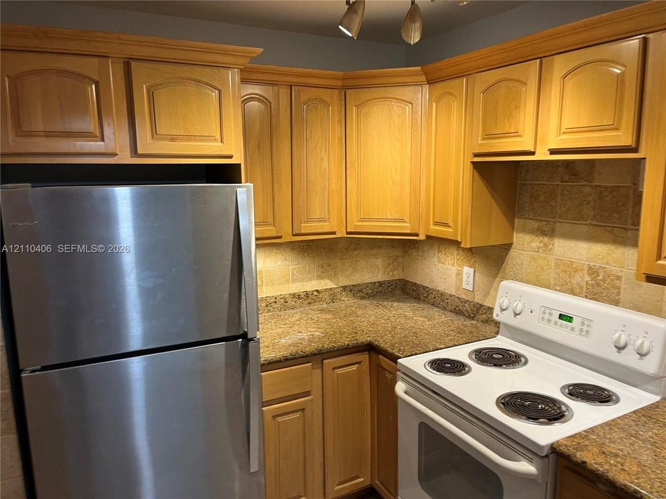 Kitchen featuring natural wood cabinetry, stone-finish countertops, square-tiled backsplash, and stainless steel refrigerator