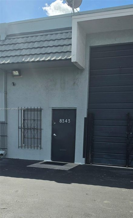 Industrial exterior featuring a high-clearance overhead roll-up door, metal-framed pedestrian entry door, security window bars, and exterior wall-mounted lighting