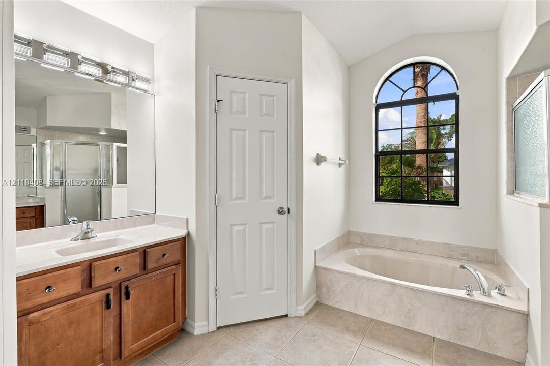 Bathroom featuring wood-finish cabinetry, stone-surround soaking tub, arched window, and large vanity mirror with integrated lighting
