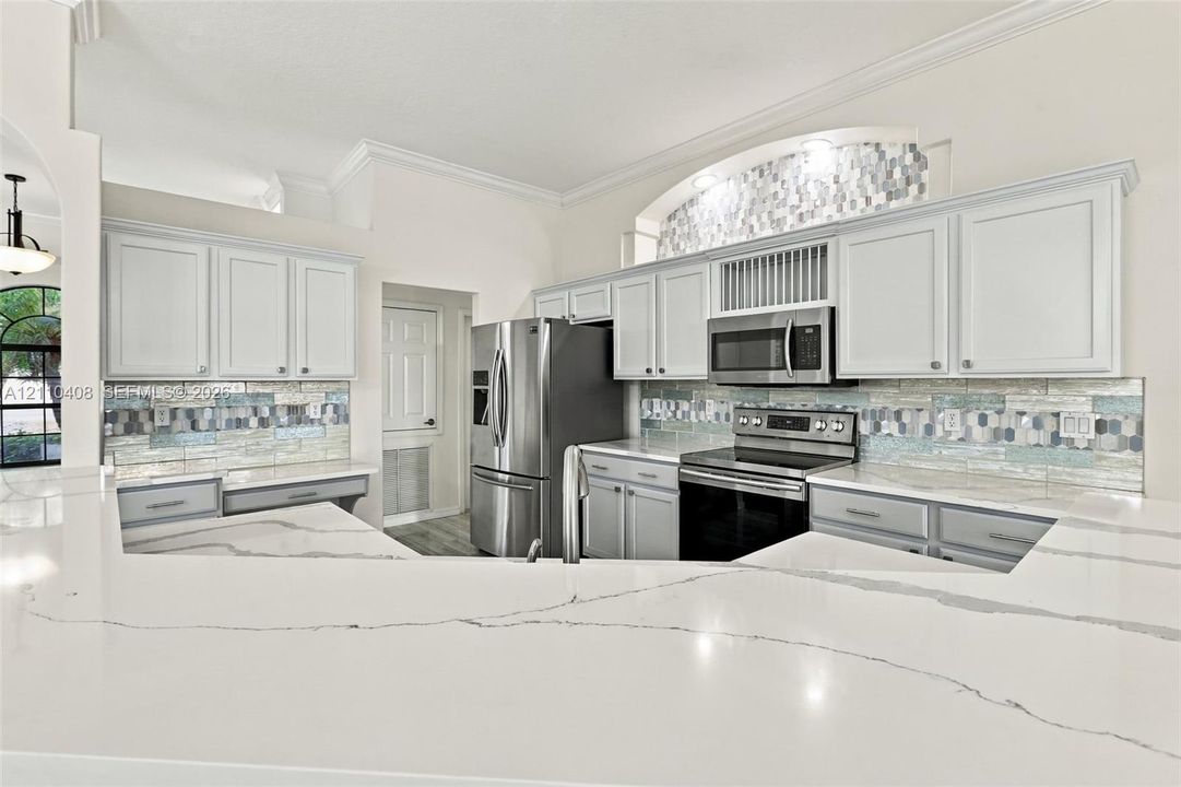 Kitchen featuring white cabinetry, solid-surface countertops with veining, stainless steel appliances, multi-toned mosaic tile backsplash, and decorative arched niche