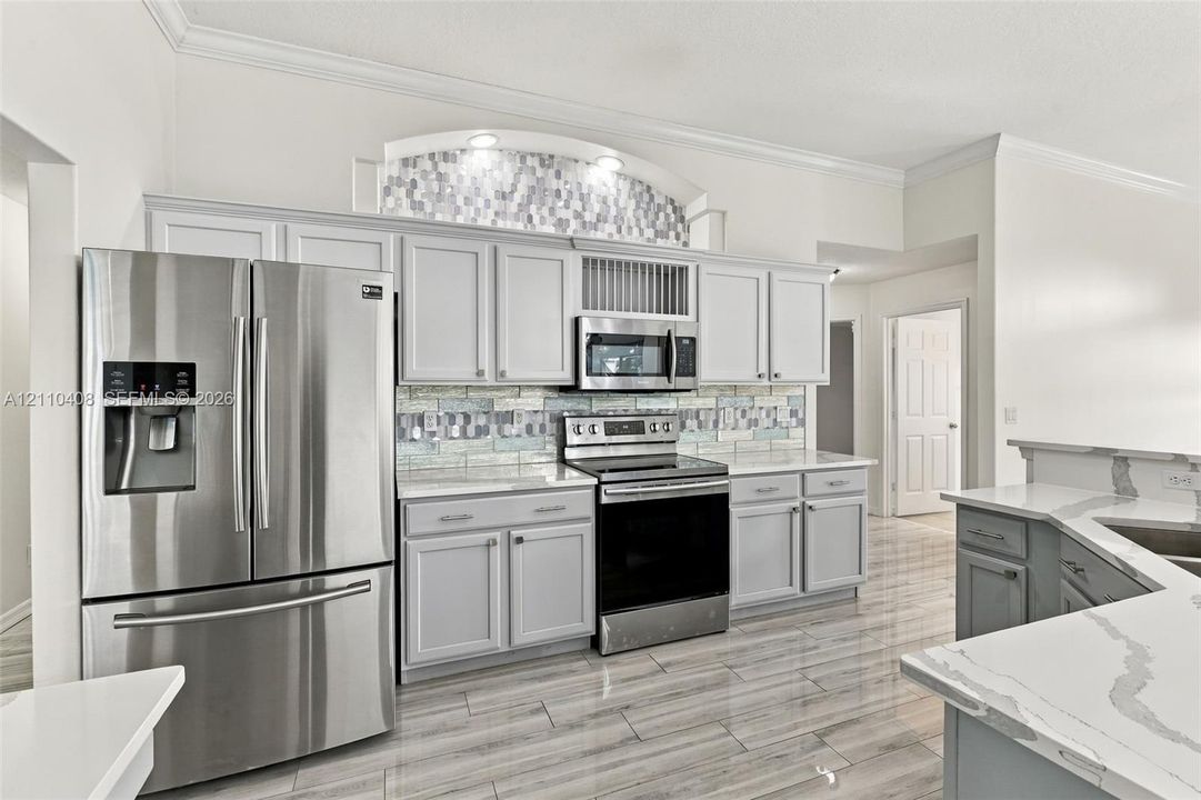 Kitchen featuring light gray cabinetry, stainless steel appliances, patterned tile backsplash, decorative arched alcove with integrated lighting, and white stone-finish countertops with veining