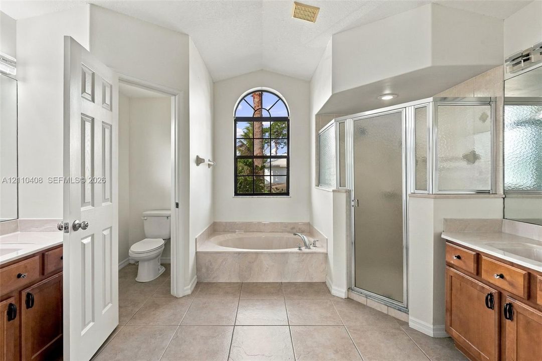 Bathroom featuring an arched window, a tiled soaking tub with a matching surround, a glass-enclosed shower, dual wood-finish vanities, and ceramic tile flooring