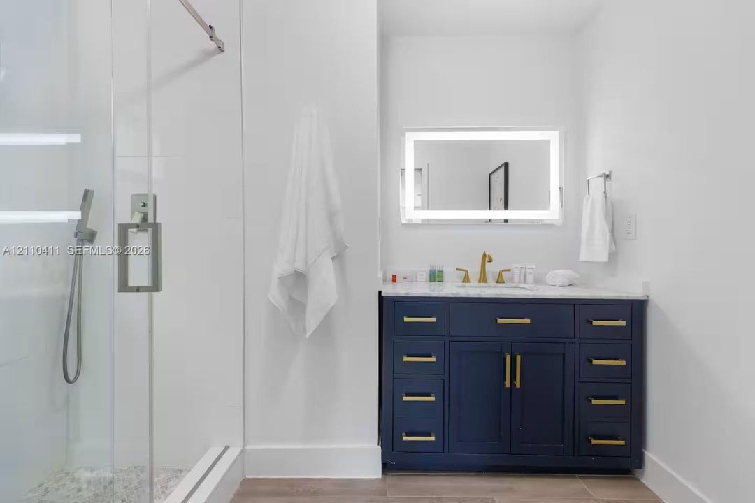 Bathroom featuring a navy blue vanity with brass-finish hardware, stone-surface countertop, frameless glass-enclosed shower with a handheld fixture, and an integrated LED-lighted mirror