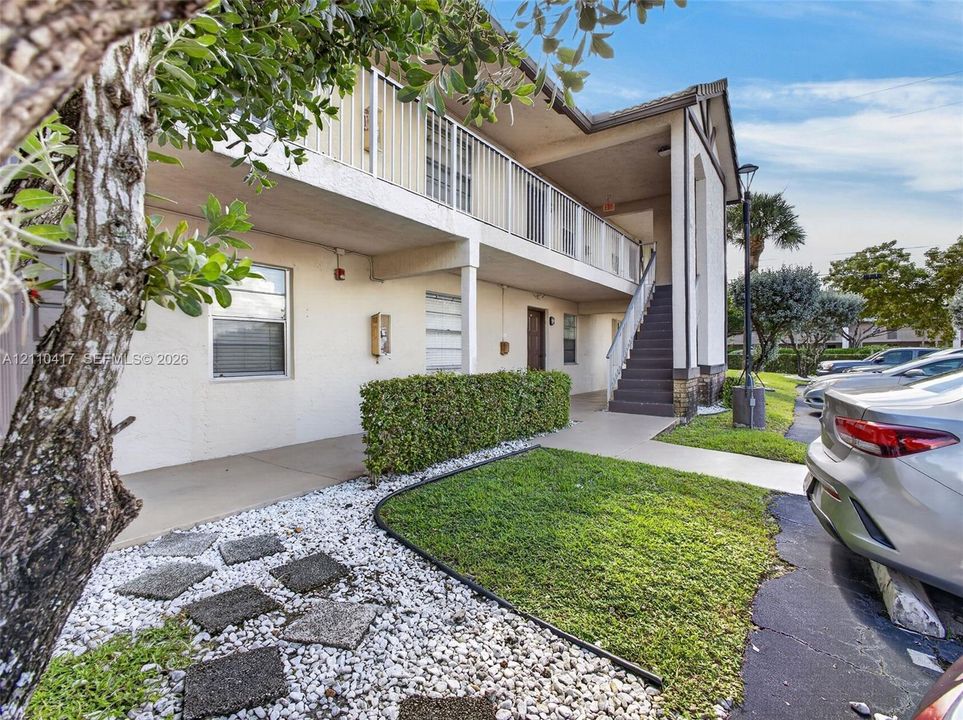 Multi-story building exterior featuring a covered balcony with metal railings, external staircase, stucco facade, and landscaped grounds with decorative stone paths