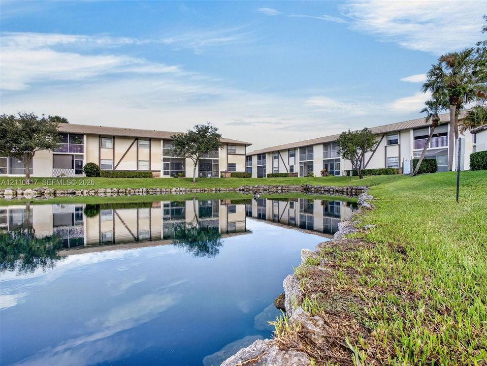 Waterfront multi-story residential building with stone-lined shore, manicured lawn, and mature palm trees