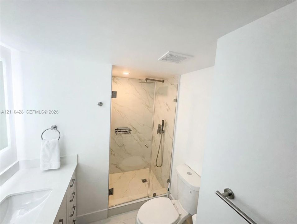Walk-in shower with stone-finish tile surround, frameless glass enclosure, rainfall shower head, handheld sprayer, and vanity with solid-surface countertop