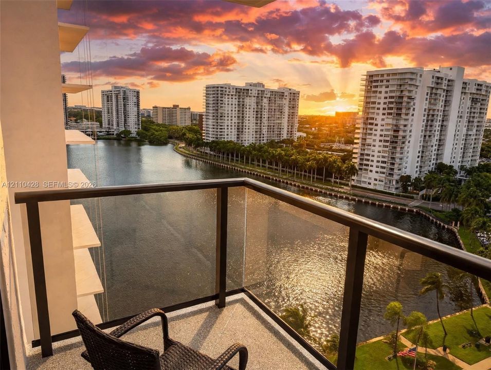 Private balcony featuring a dark metal railing with integrated glass panels, textured concrete flooring, and a backdrop of waterfront scenery at sunset
