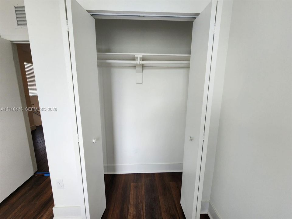 Reach-in closet featuring white dual-panel doors, an integrated upper storage shelf, and a hanging rod, complemented by dark wood-finish flooring