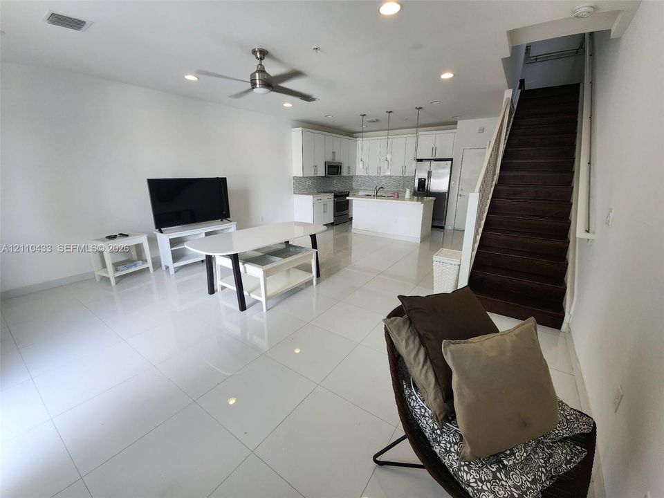 Open-concept interior featuring large-format tile flooring, white cabinetry, a kitchen island, recessed lighting, and a staircase with dark wood-finish treads