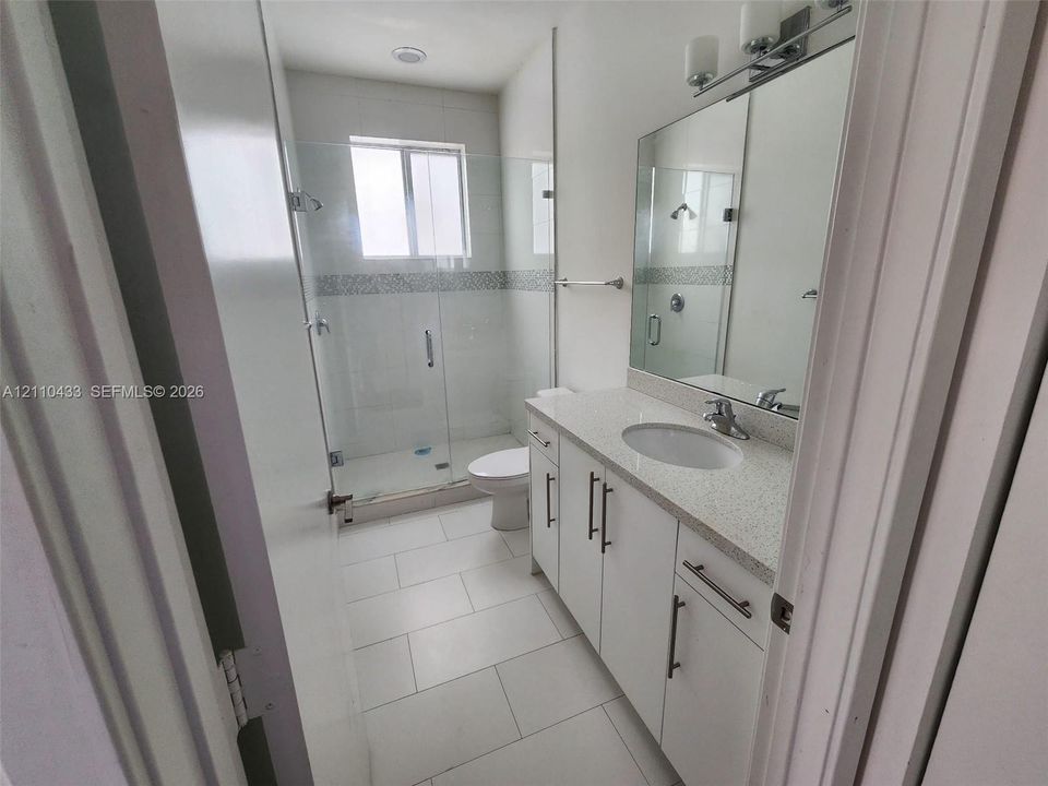 Bathroom featuring white cabinetry, light-toned stone-finish countertop, walk-in shower with a glass enclosure and decorative tile inlay, and large-format tile flooring