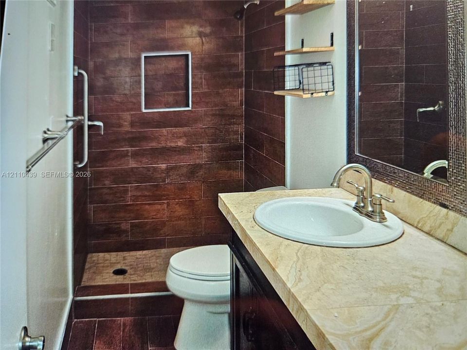 Walk-in shower featuring dark wood-finish tile walls and a built-in recessed niche, stone-finish vanity countertop with an undermount sink, and wall-mounted shelving