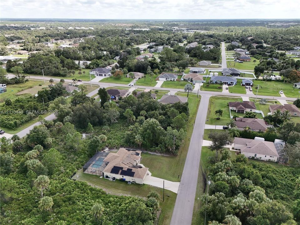 Aerial perspective featuring a single-story residence with an attached screened-in pool enclosure, integrated solar panels, and an asphalt shingle roof, positioned on a lot surrounded by mature native landscaping