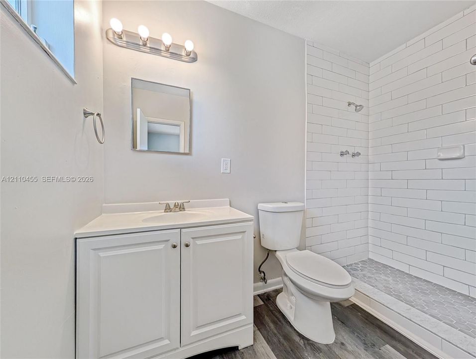 Bathroom featuring white shaker-style cabinetry, a molded vanity countertop with a chrome faucet, wood-finish flooring, and a walk-in shower with white subway-style tile surround and hexagonal floor tile