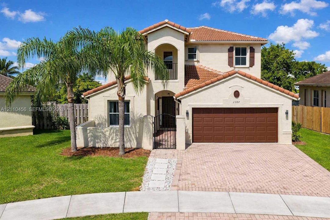 Two-story stucco exterior featuring a tile roof, second-story balcony, attached two-car garage, and paver driveway
