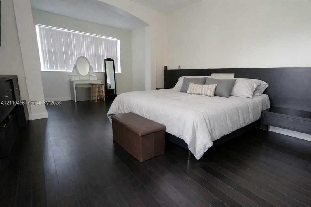 Room featuring dark wood-finish flooring, a built-in dark-toned headboard with integrated side tables, an arched architectural alcove, and floor-to-ceiling windows with vertical blinds