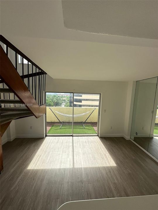 Open-concept interior featuring wood-finish flooring, a metal staircase with wooden handrail, expansive mirrored wall panels, and sliding glass doors opening to a private balcony with synthetic turf