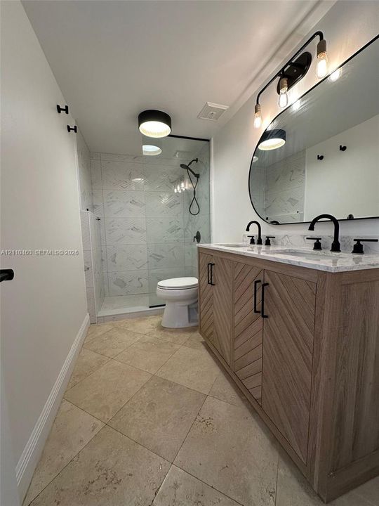 Double vanity featuring wood-finish chevron cabinetry, stone-style countertop, matte black fixtures, and a walk-in shower with floor-to-ceiling tile