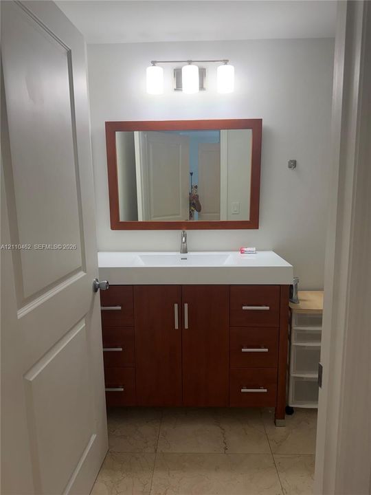 Vanity featuring wood-finish cabinetry, a rectangular white sink, a solid surface countertop, a matching wood-frame mirror, and a three-light fixture