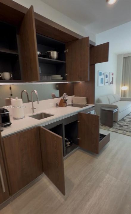 Custom kitchenette featuring dark wood-finish cabinetry, quartz countertops, a stainless steel undermount sink, integrated under-cabinet lighting, and a mirrored backsplash