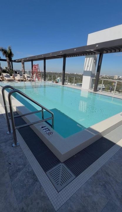 Rooftop swimming pool featuring a tile border, metal guardrails, a modern pergola structure, and glass perimeter fencing