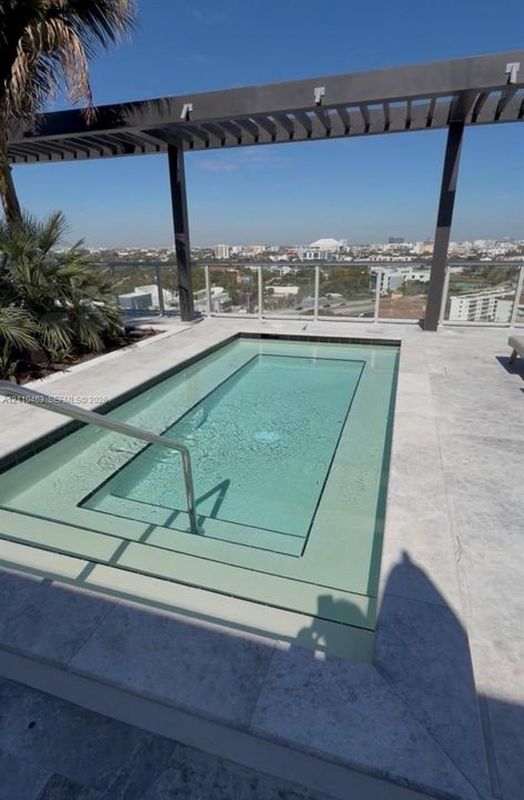 Rooftop plunge pool featuring natural stone tiling, a metal pergola, a glass panel railing, and integrated metal handrail