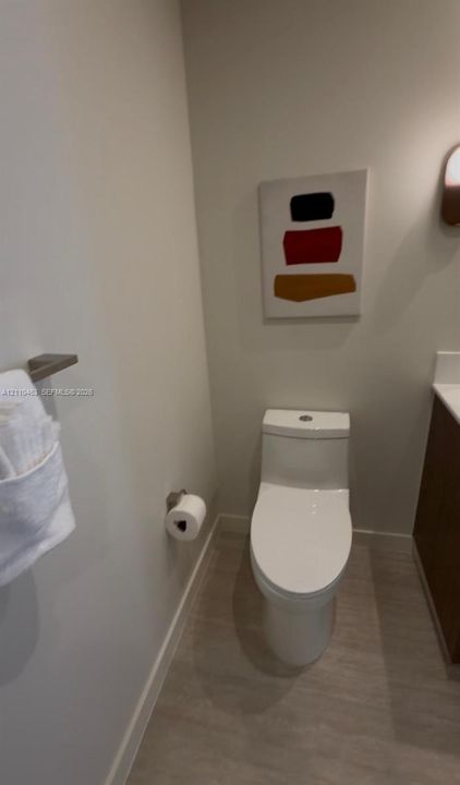 Powder room featuring a dual-flush elongated toilet, wood-finish flooring, wall-mounted brushed nickel towel bar, and a wall-mounted light fixture