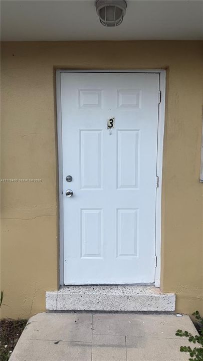 White six-panel entry door featuring a circular hardware set, deadbolt, and mounted overhead light fixture, finished with a concrete doorstep