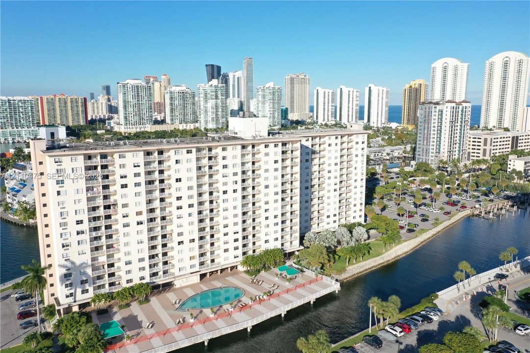 High-rise residential building featuring exterior balconies, a ground-level swimming pool, and concrete hardscaping situated along a waterway