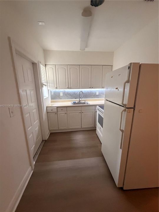 Kitchen featuring white raised-panel cabinetry, light-toned countertops, under-cabinet lighting, a stainless steel sink with a gooseneck faucet, and wood-finish flooring