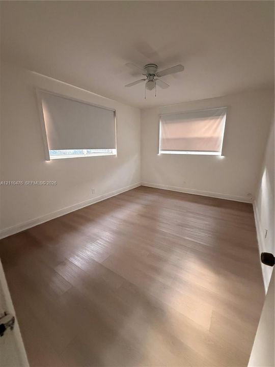 Room featuring wood-finish flooring, white walls, dual windows with roller shades, and a ceiling fan