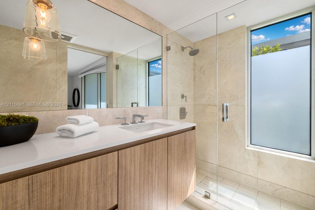 Wood-finish floating vanity with white solid surface countertop, undermount sink, glass-enclosed shower with full-height stone-finish wall tile, and large wall-to-wall mirror