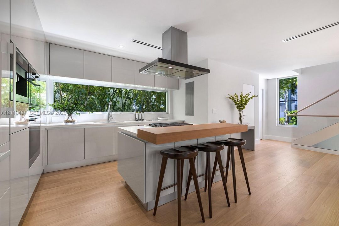 Modern kitchen featuring gloss-finish cabinetry, wood-finish flooring, a central island with a wood-tone countertop, a stainless steel range hood, and a horizontal window backsplash