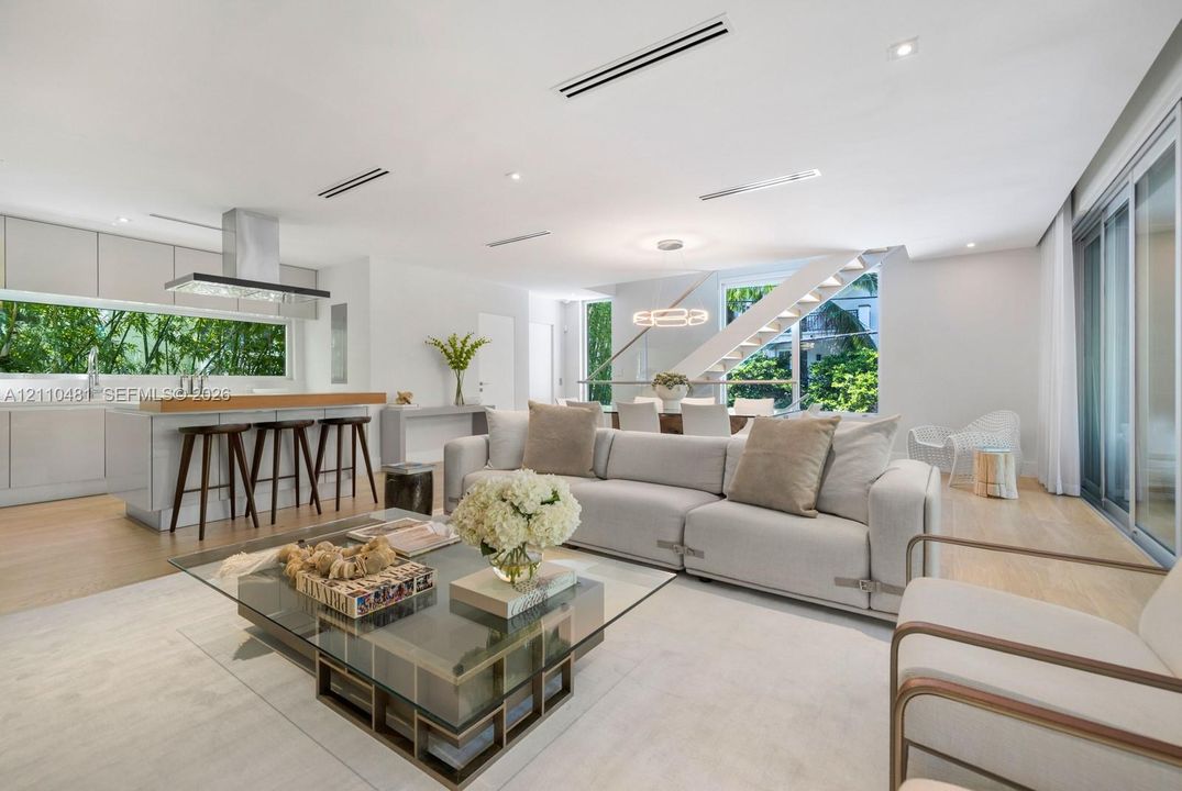 Open-concept interior featuring light wood-finish flooring, linear recessed lighting, a modern floating staircase with glass railings, large horizontal windows, and a stainless steel kitchen ventilation hood