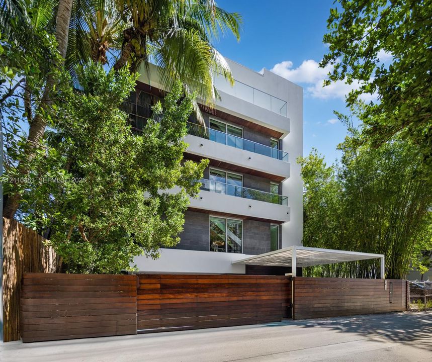 Modern multi-story exterior featuring white stucco and dark stone cladding, stacked balconies with glass railings, and a horizontal wood slat perimeter fence