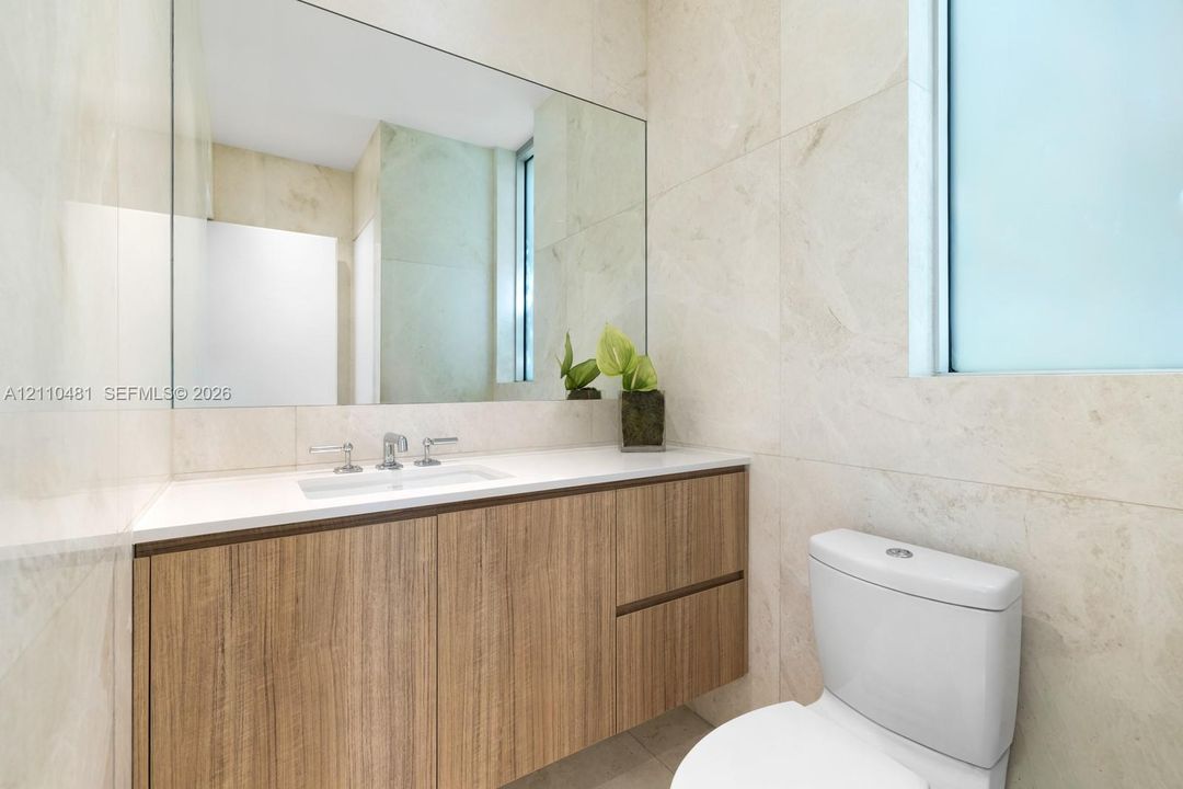 Wall-mounted vanity featuring wood-finish cabinetry, white countertop with integrated sink, chrome widespread faucet, large frameless mirror, and floor-to-ceiling neutral tile work