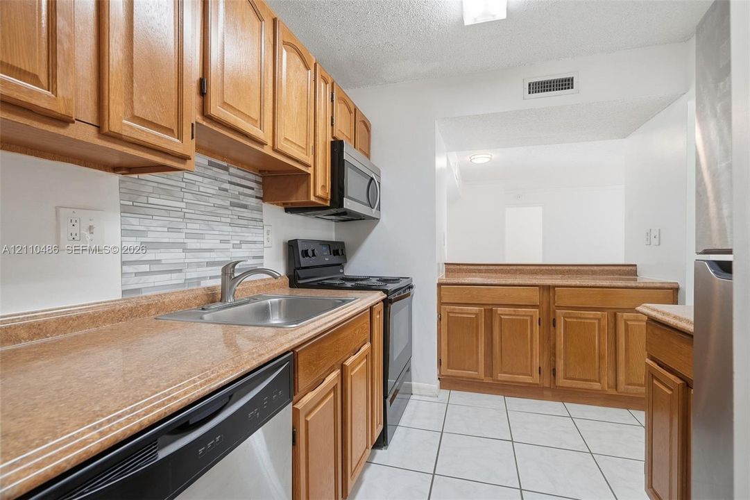 Kitchen featuring wood cabinetry, stainless steel appliances, solid-surface countertops, decorative stone-finish backsplash, and tile flooring