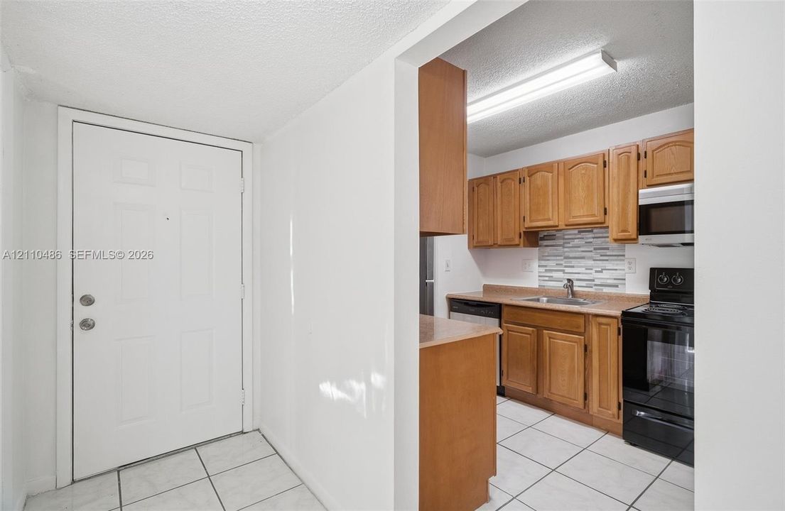 Kitchen featuring wood cabinetry, tiled backsplash, solid surface countertops, and black appliance suite, complemented by white tile flooring