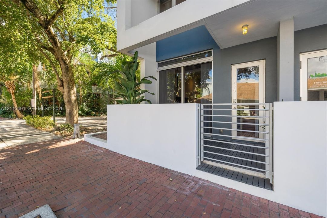 Modern exterior entrance featuring a white stucco privacy wall, horizontal slat metal gate, red brick paver walkway, and recessed overhead lighting
