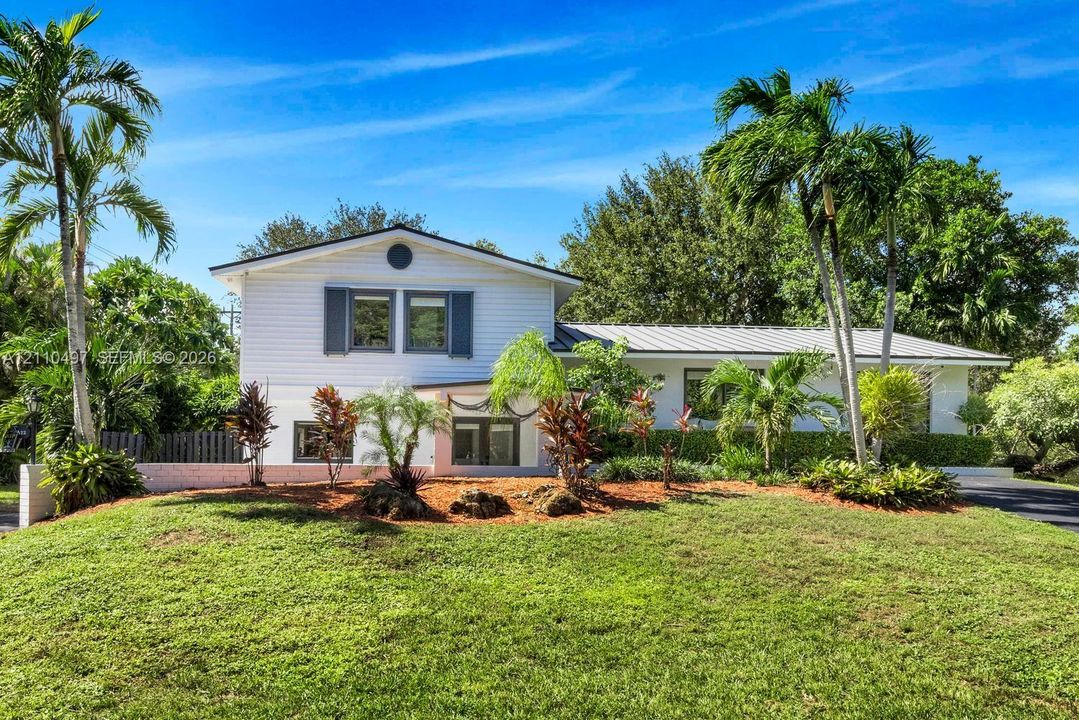 White siding exterior featuring a standing seam metal roof, shuttered windows, and tiered landscaping with mature palm trees
