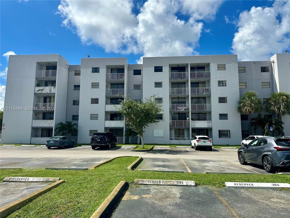Mid-rise building exterior featuring a multi-story facade, private balconies with metal railings, and a paved surface parking lot