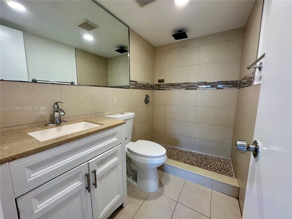 White vanity with a stone-finish countertop, undermount rectangular sink, polished chrome faucet, tile-surround shower with a pebble-tile floor accent, and large wall-mounted mirror