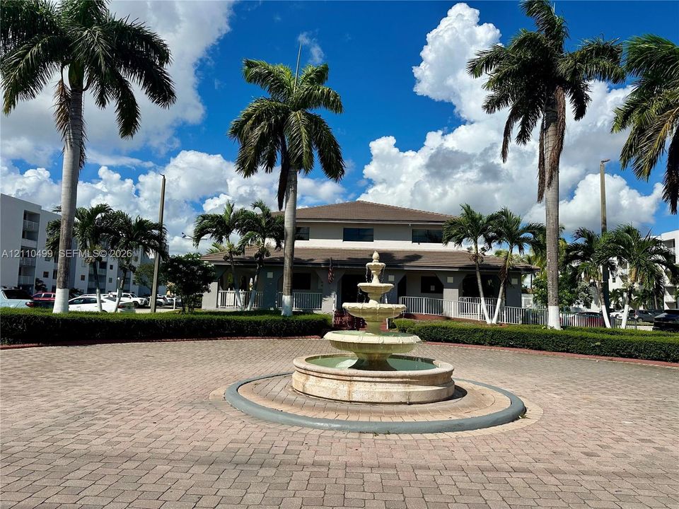 Circular paver courtyard featuring a multi-tiered stone fountain, mature palm trees, and manicured shrubbery