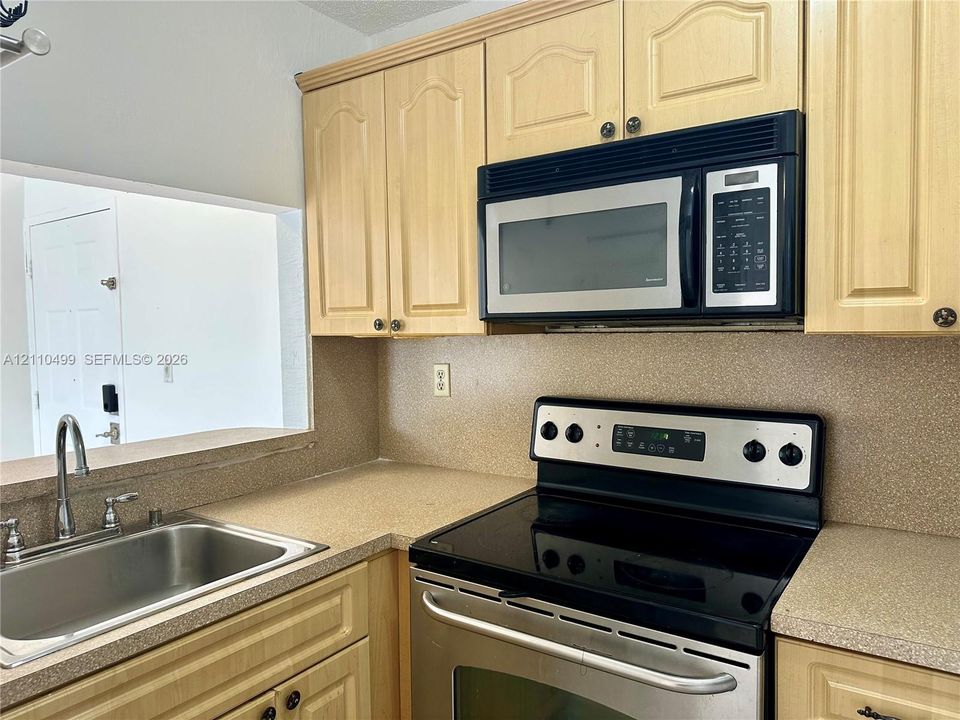 Kitchen featuring light-finish cabinetry, stainless steel microwave, stainless steel range with electric glass cooktop, solid-surface countertops, and a stainless steel sink with a high-arch faucet