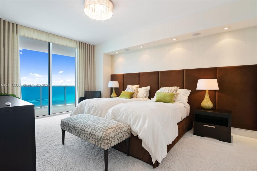 Floor-to-ceiling glass sliding doors opening to a balcony, custom brown upholstered headboard wall, recessed ceiling lighting, and a drum-style chandelier