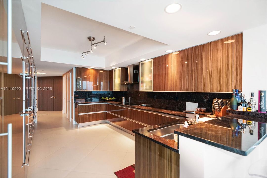 Gourmet kitchen featuring high-gloss wood-finish cabinetry, dark stone countertops, stainless steel appliances, recessed ceiling lighting, and large-format tile flooring