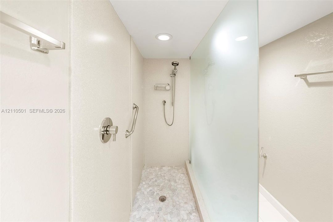 Walk-in shower featuring a frosted glass panel, pebble-stone flooring, brushed-nickel handheld shower head, and wall-mounted metal support bars