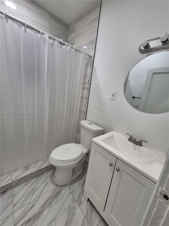 Bathroom featuring large-format marbled tile flooring, a white vanity cabinet with an integrated basin, a round wall-mounted mirror, and a tiled shower surround